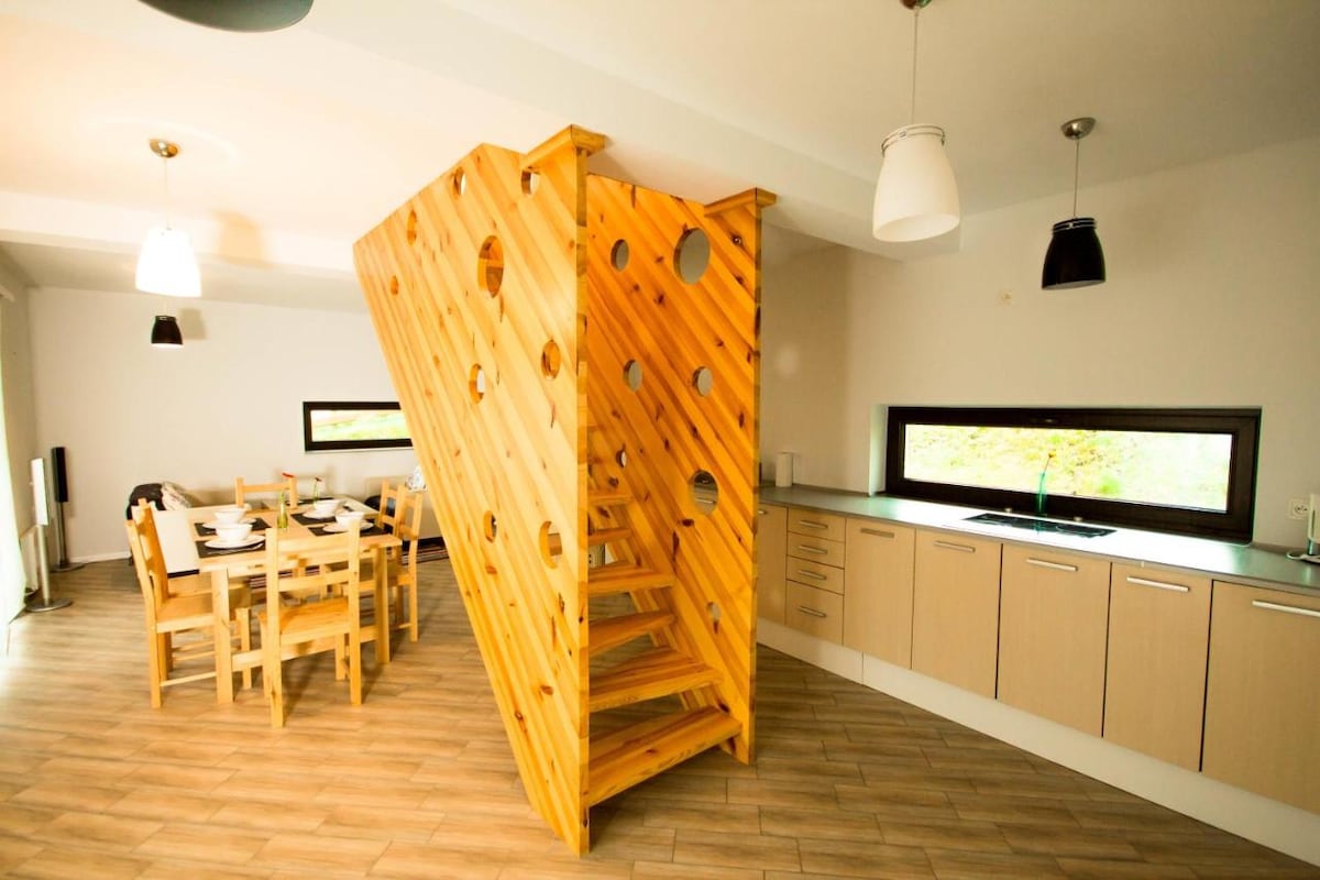 A unique wooden staircase with circular cutouts is centrally featured in an open dining area. Surrounding it are light wooden chairs and a modern kitchen with beige cabinetry. Large windows allow natural light to fill the space, creating a bright and airy environment.
