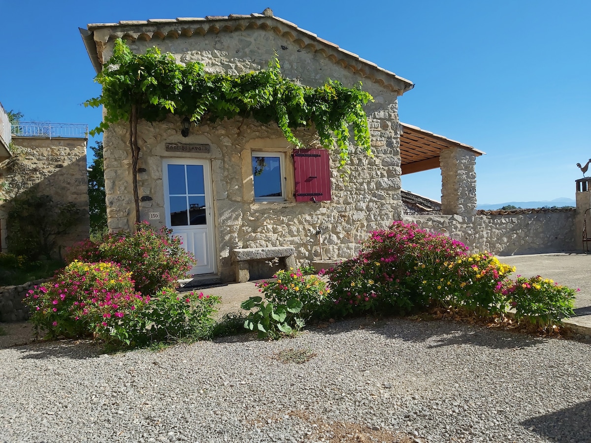The charming stone cottage is framed by vibrant flowering shrubs and lush greenery. A welcoming entrance features a white door and pink shutters. A covered outdoor area provides shade, while the clear blue sky serves as a backdrop, enhancing the serene outdoor space.