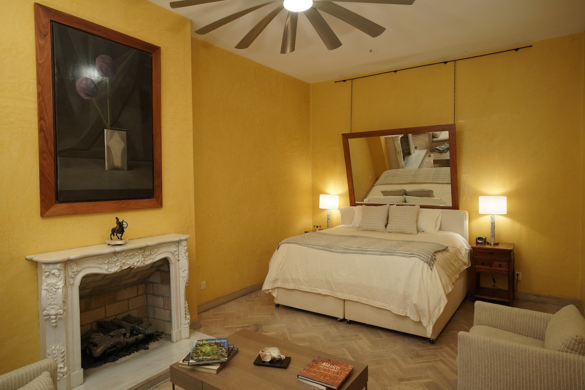 A spacious bedroom features warm yellow walls and a large mirror above the bed, reflecting natural light. A white bed with soft linens is complemented by side tables and lamps. A cozy seating area includes light-colored chairs, and a decorative fireplace is present.