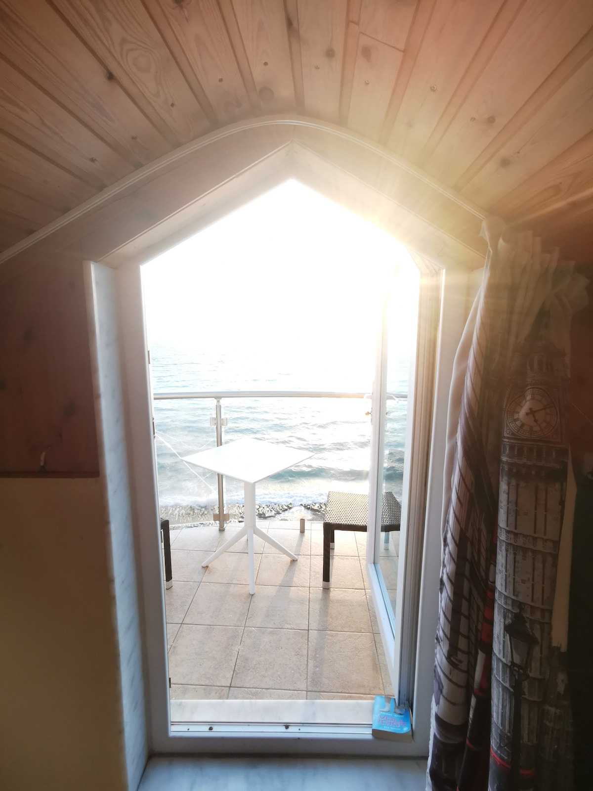 A balcony door opens to reveal a view of the sea, with sunlight streaming in from the outside. A small table and two chairs are positioned on the tiled patio, inviting moments of relaxation by the water.