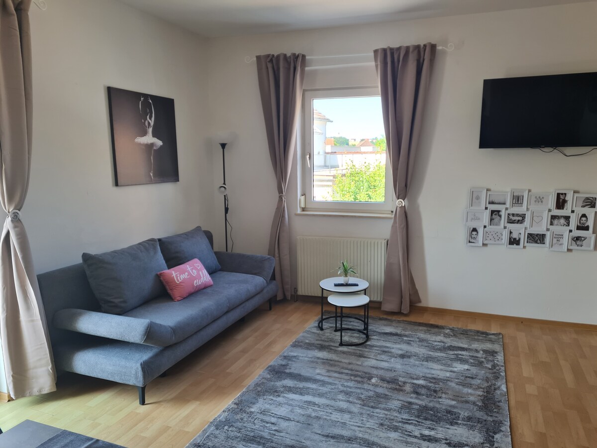 A modern living area features a gray sofa with a decorative pillow. A small white table is positioned in front of the sofa. Large windows allow natural light to fill the space, adorned with light-colored curtains. A flat-screen TV is mounted on the wall opposite.