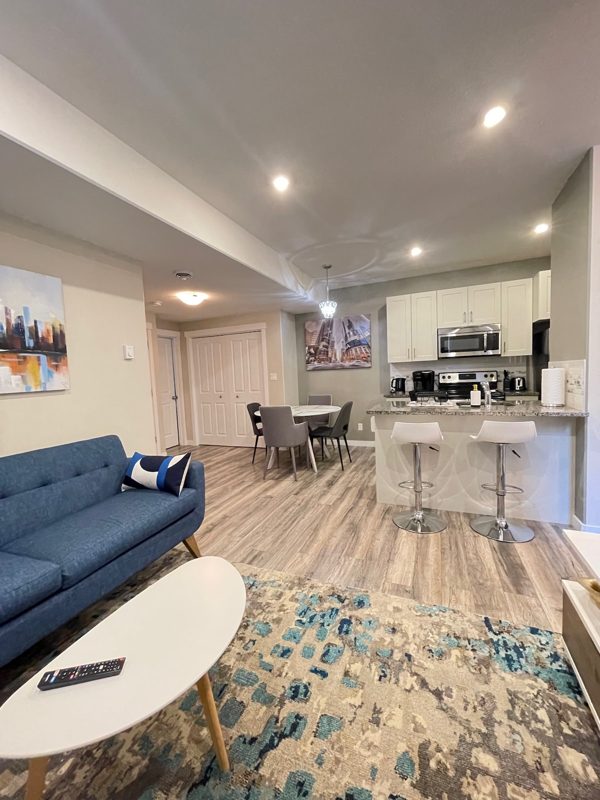 A modern living space features a blue sofa, a round coffee table, and a patterned area rug. The dining area includes a table with four chairs, while the kitchen is visible in the background, showcasing stainless steel appliances and quartz countertops.
