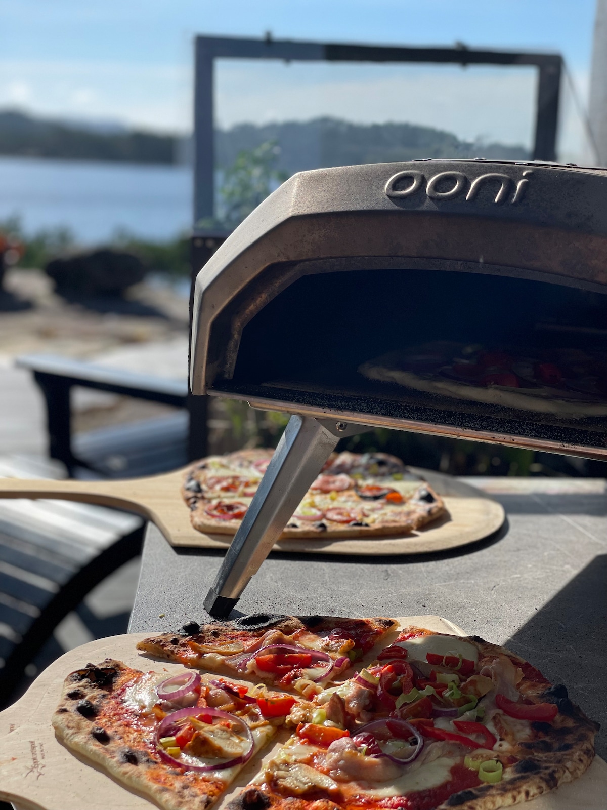An outdoor pizza oven is featured prominently, with freshly baked pizzas placed on wooden peels. The table is set against a backdrop of a serene body of water and distant hills, under a clear sky. Seating is visible nearby.