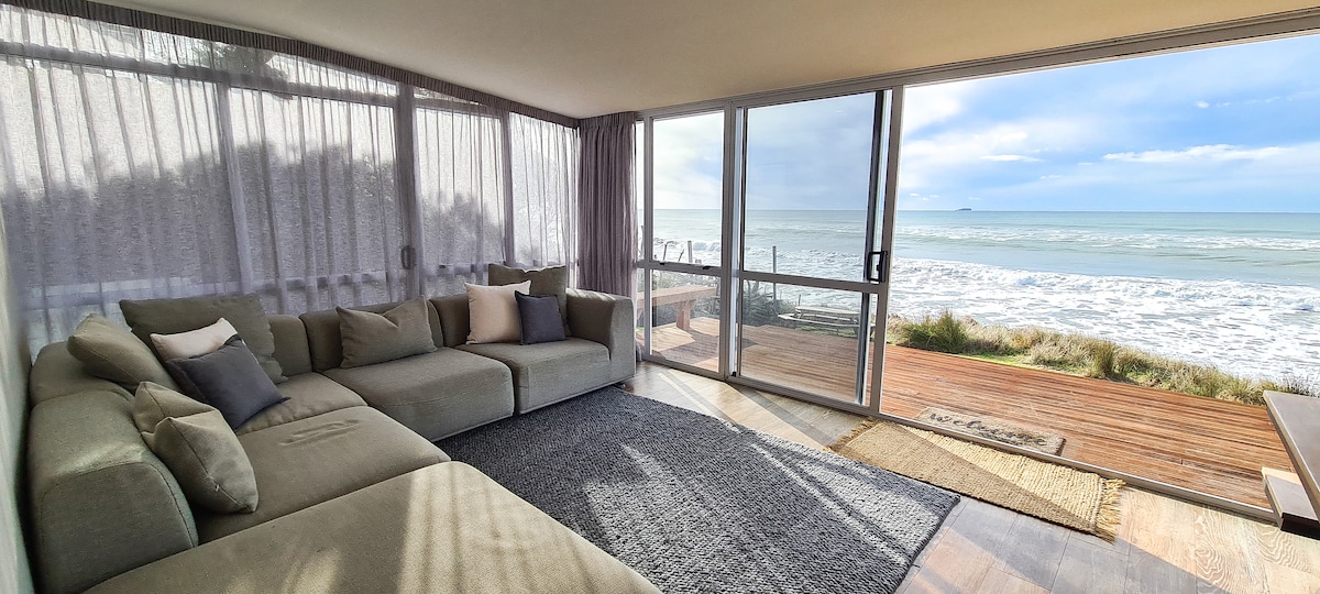 A spacious lounge area features a large sectional sofa with soft cushions. Sheer curtains allow natural light to fill the room, showcasing panoramic ocean views through expansive glass doors that open to the deck facing the beach.