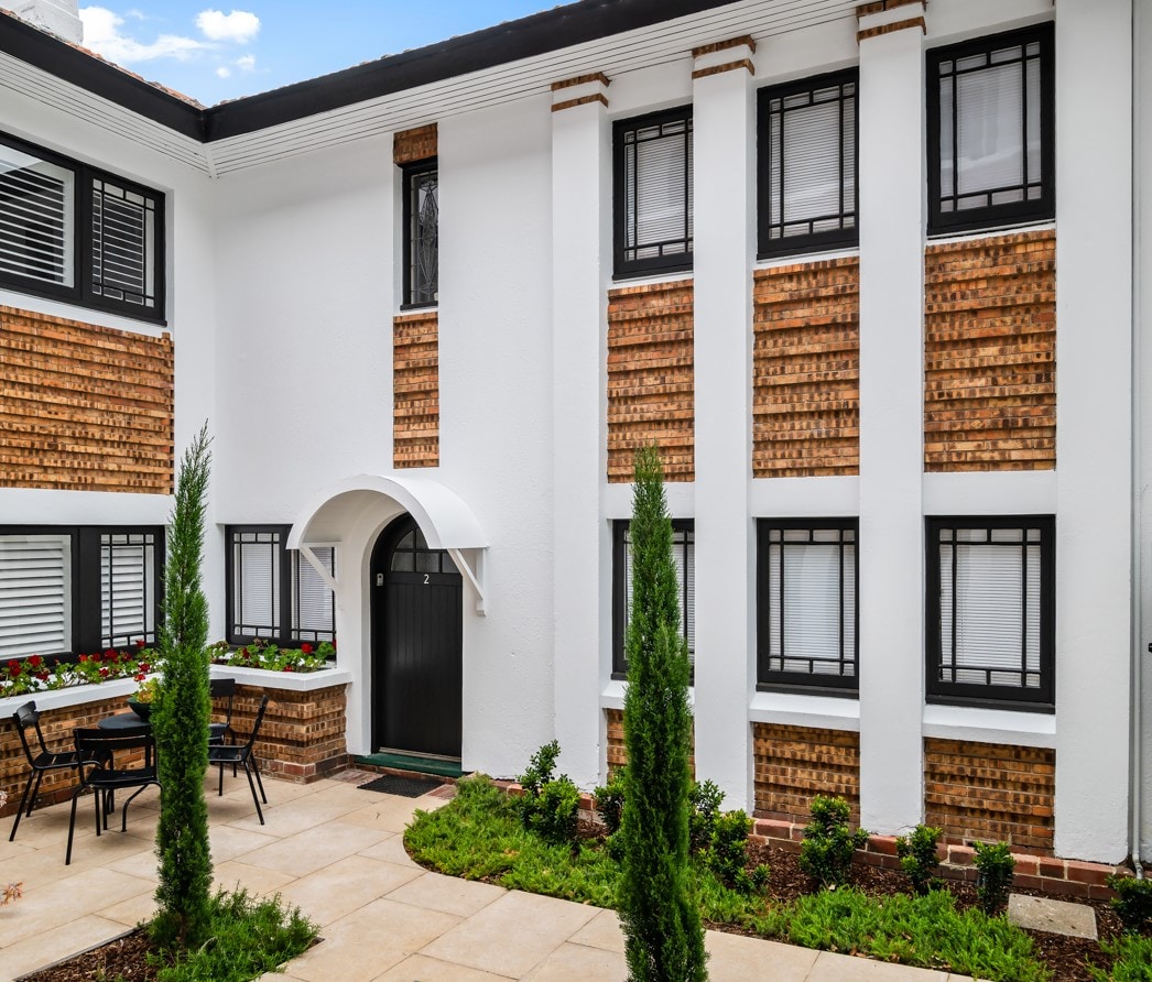 The exterior of the heritage apartment showcases a combination of white and textured brown brick walls. A private courtyard area features a table and chairs set against a backdrop of neatly arranged greenery and decorative plants.