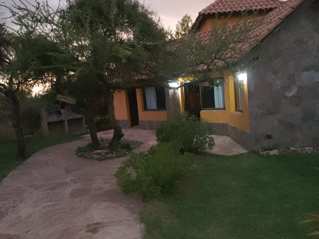 A stone cabin is set within a grassy landscape, featuring large windows that frame the entrance. A winding path leads to the front door, with trees providing natural shading. Soft lighting from the exterior accents the warm hues of the walls and roof.