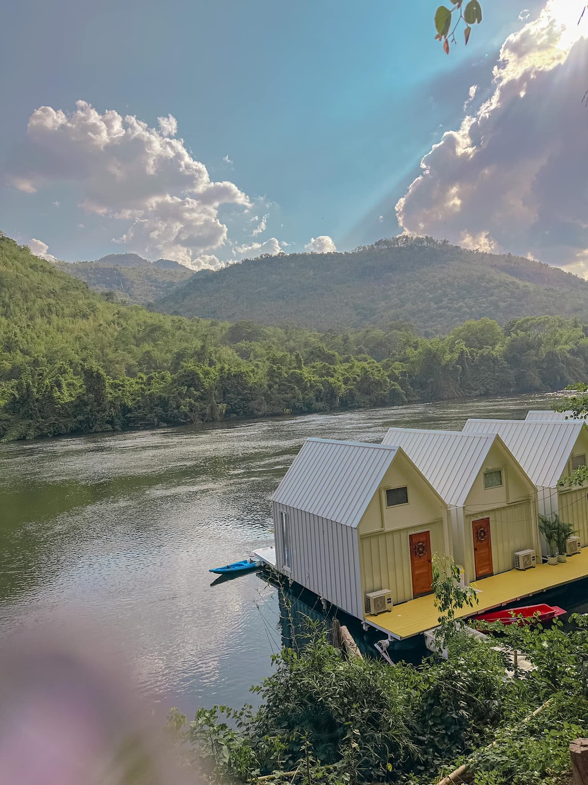 Two houseboats are positioned along the serene riverbank, surrounded by lush green hills and gentle water. The mountains rise in the background under a partly cloudy sky, with sunlight filtering through, creating a peaceful atmosphere. A kayak is visible near the shore.