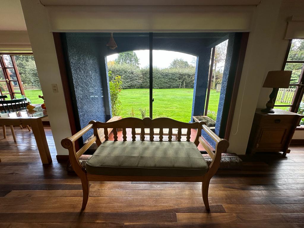 A wooden bench with green cushions is positioned indoors, facing a large sliding glass door. The door opens to a lush green garden, framed by dark blue walls. Natural light brightens the space, highlighting the warm wooden flooring.