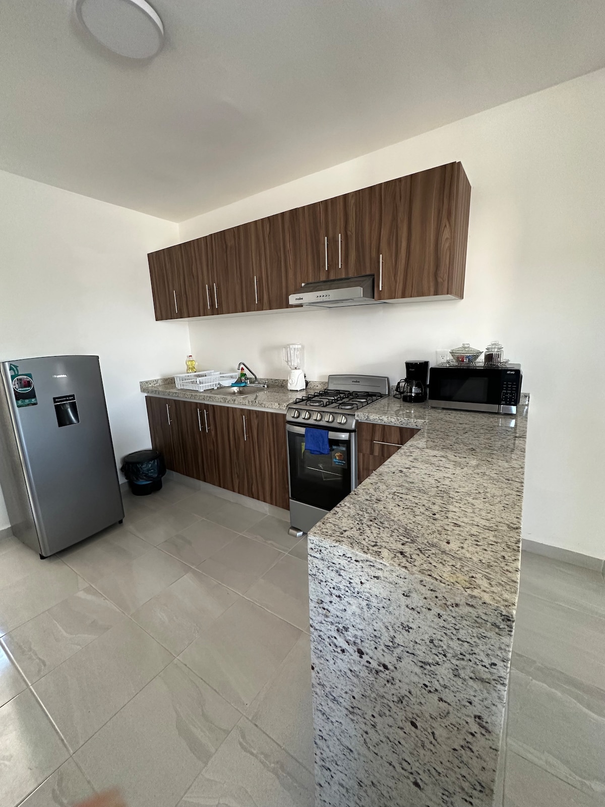A modern kitchen features sleek wooden cabinets and a spacious countertop. Appliances include a refrigerator, gas stove, and microwave. The area is well-lit with natural light, highlighting the polished tile flooring and organized setup for cooking and dining.