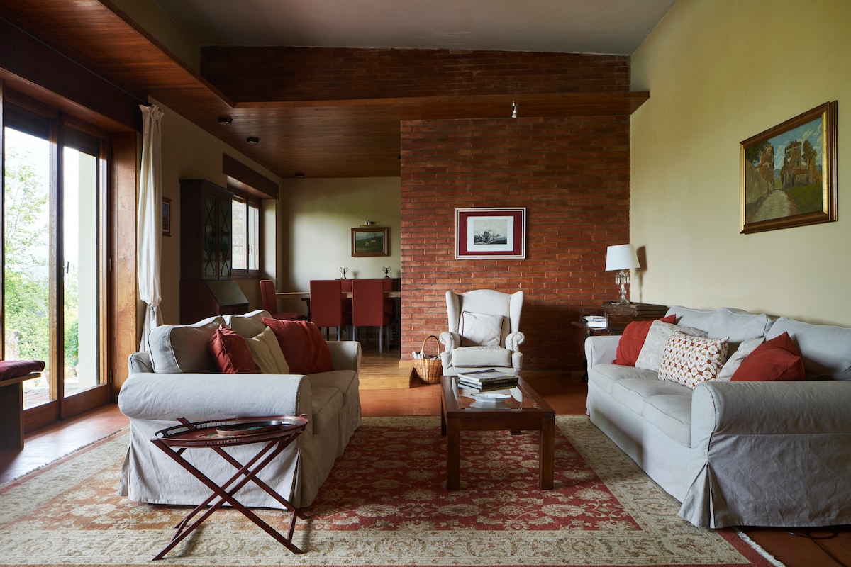 A spacious living room is furnished with light-colored sofas adorned with colorful cushions. A coffee table sits in the center, surrounded by elegant wooden accents. Large glass doors allow natural light to enter and provide views into the dining area beyond.