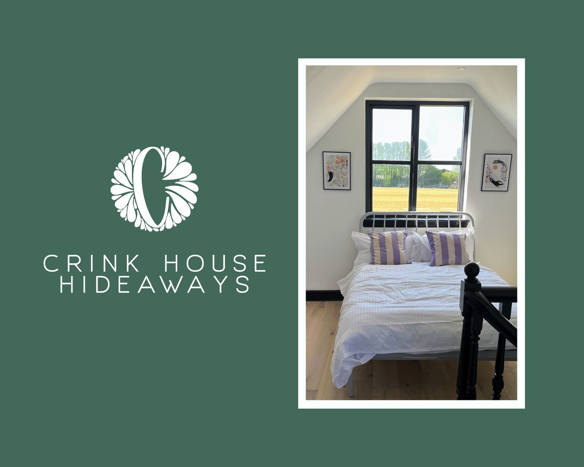 A bright sleeping area features a bed with white linens and purple striped pillows. Two framed artworks adorn the walls beside the window, which offers views of the open fields beyond. The room is completed with light wooden flooring and a decorative black railing.