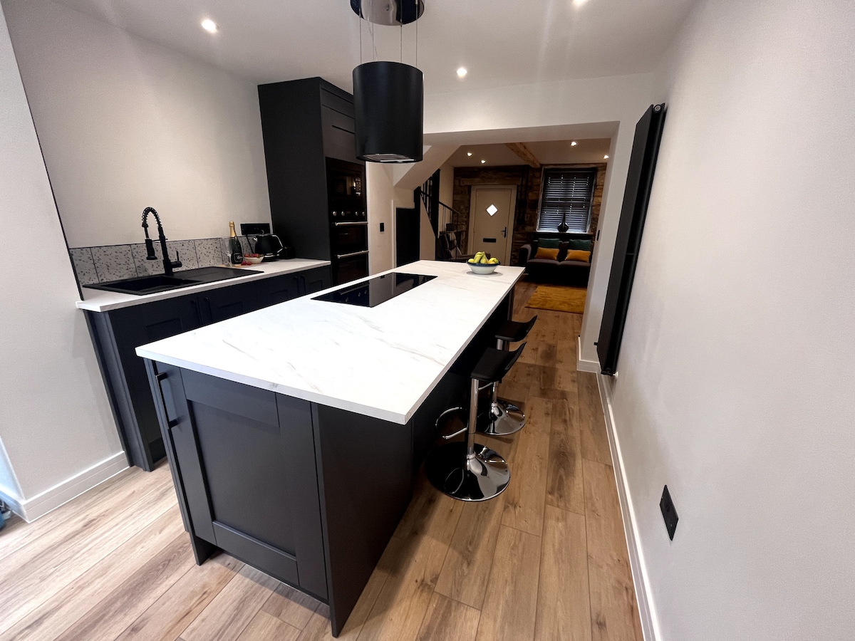 A modern kitchen features a sleek black cabinetry and a spacious white marble island with seating for four. An induction cooktop is embedded in the island, complemented by minimalist lighting and natural wood flooring that enhances the contemporary design.