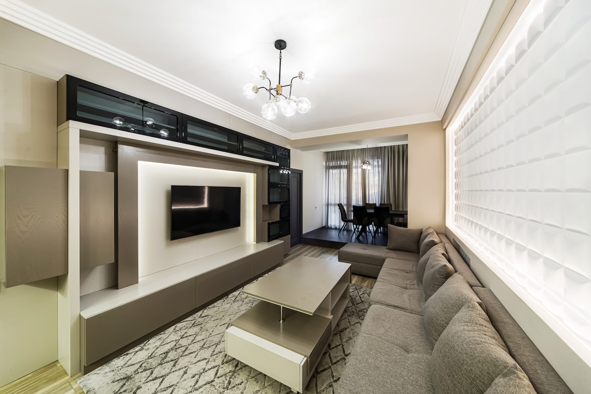 A well-lit living area features a modern sectional sofa in neutral tones, paired with a sleek coffee table. A wall-mounted television is integrated within a stylish cabinet. Natural light enters through sheer curtains, and a dining table with chairs is visible in the background.