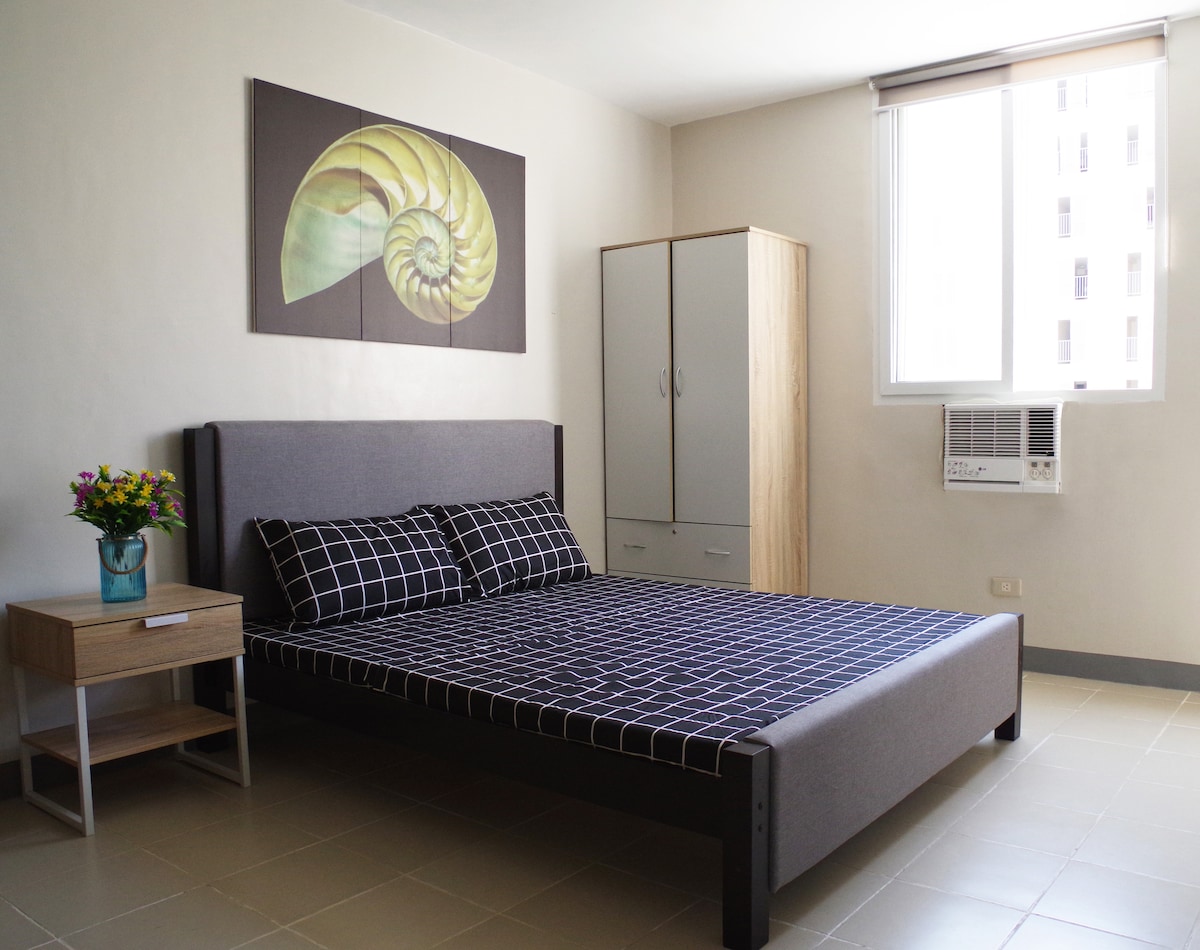 A simple yet stylish bedroom features a large bed with a dark patterned cover, complemented by two matching pillows. A modern painting of a spiral shell adorns the wall above the bed. Natural light filters through a nearby window, while a cabinet and bedside table complete the space.