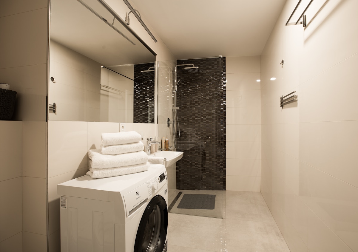 A modern bathroom is presented, featuring a spacious shower with a sleek black tile backdrop. A washing machine is positioned conveniently with neatly stacked white towels on top. Soft lighting enhances the clean, minimalist design, emphasizing the bright, neutral tones throughout the space.