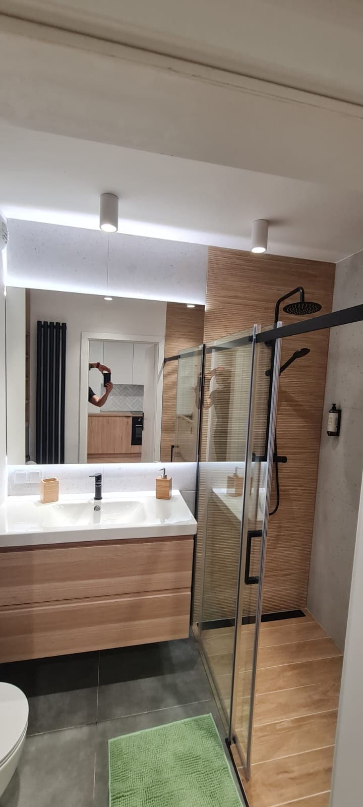 A modern bathroom features a spacious shower enclosed in glass, with sleek fixtures mounted on a wooden accent wall. A vanity with a crisp white sink is complemented by natural wood cabinetry. Subtle lighting enhances the neutral tones throughout the space.