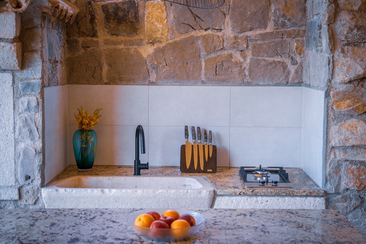 A rustic outdoor kitchen is depicted, featuring a stone wall and granite countertop. A stylish black faucet is positioned above a deep sink. A wooden knife block with several knives adds functionality, while a gas stove is visible on the granite surface. A bowl of fresh fruit is placed in the foreground.