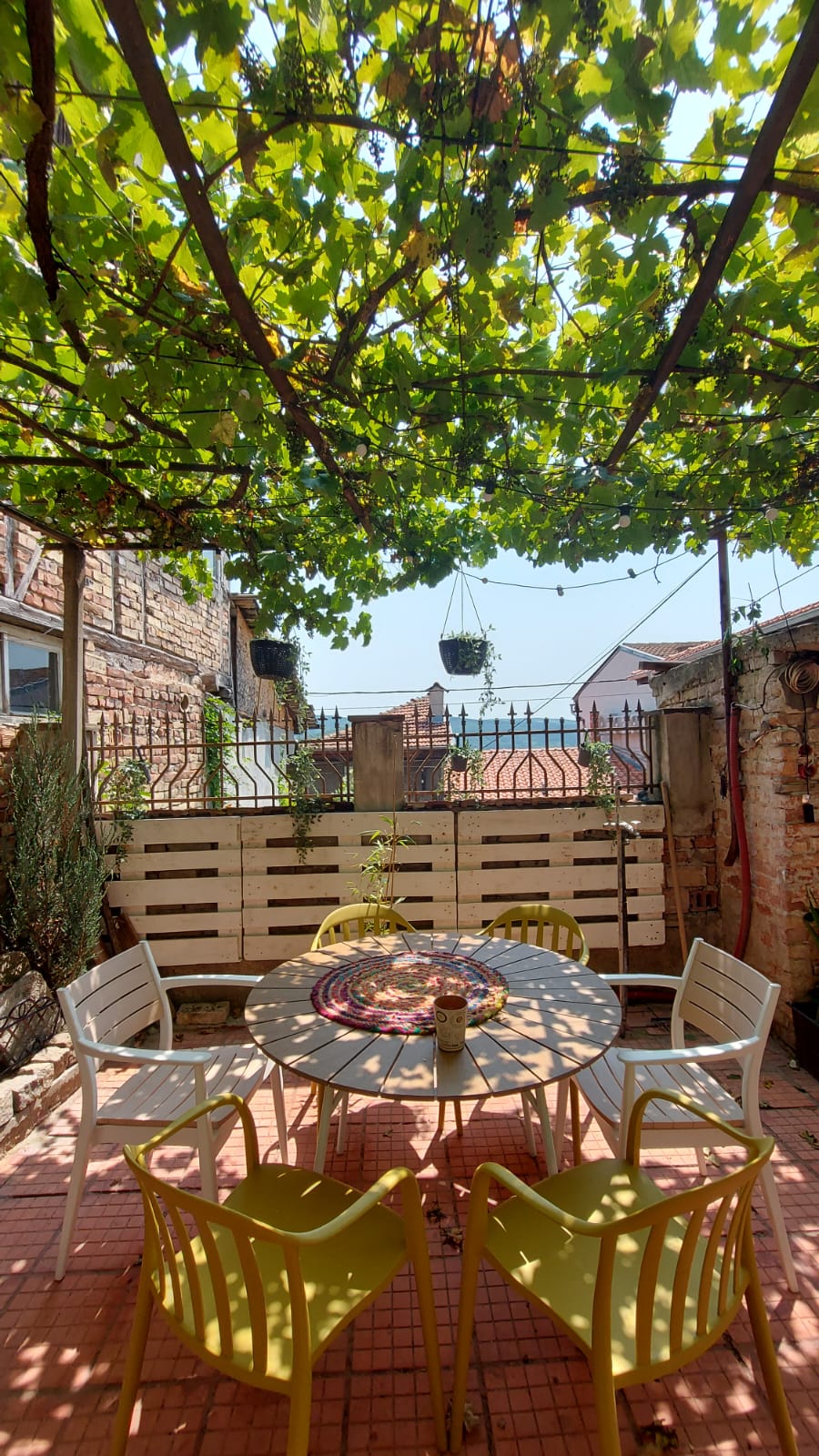 A private outdoor space features a round table surrounded by colorful chairs, nestled under a canopy of lush greenery. The setting allows for relaxed dining or socializing, with decorative hanging plants and a fence providing privacy while maintaining an inviting ambiance.