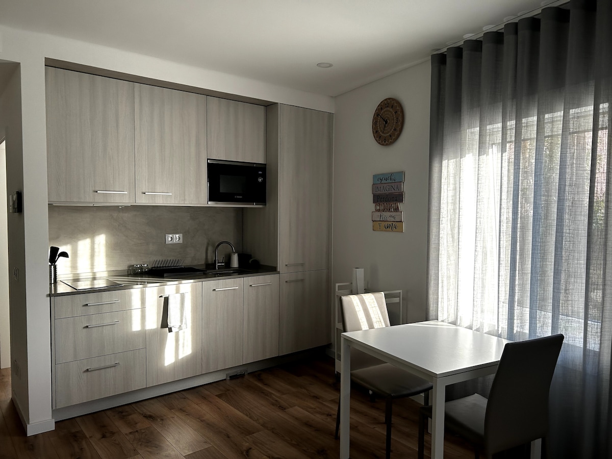 A modern kitchen is visible, featuring light wood cabinets and stainless steel appliances, including a microwave. A white dining table with two chairs is set beside a large window, dressed in sheer gray curtains that allow natural light to filter in.