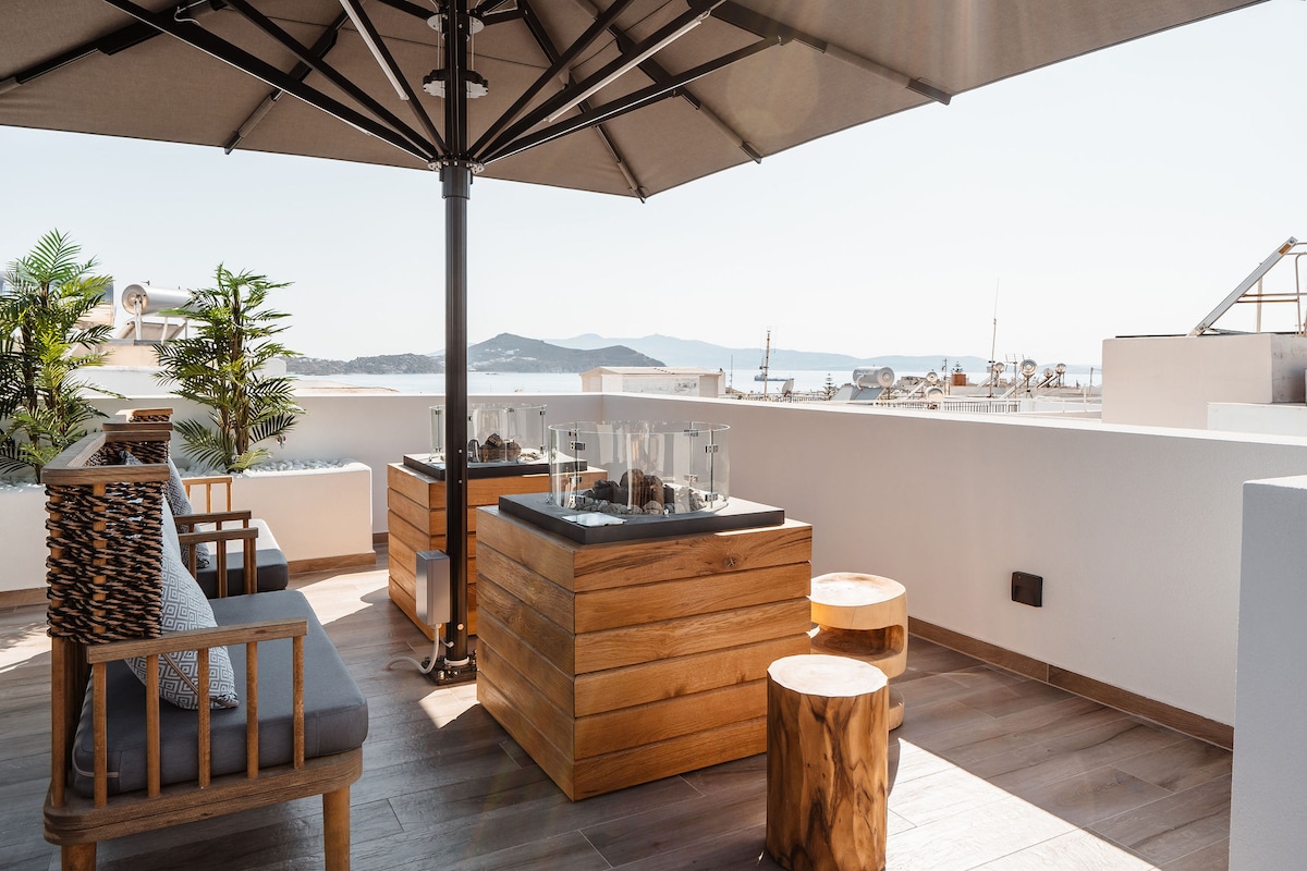 A rooftop garden area features comfortable seating with soft cushions under a large canopy. Natural wood elements, including a fire pit and small tables, complement the space. Scenic sea views and distant mountains are visible on the horizon, enhancing the serene atmosphere.