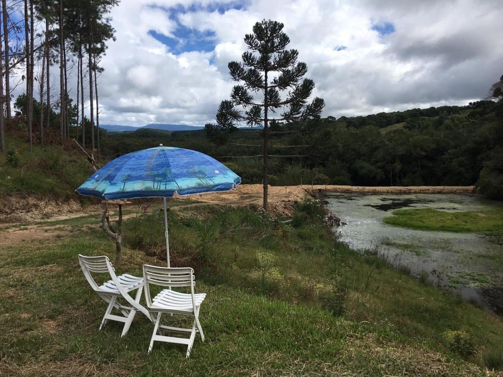 Two white lounge chairs are positioned on the grass near a small pond, accompanied by a blue umbrella providing shade. Surrounding trees and verdant hills are visible in the background, under a partly cloudy sky.