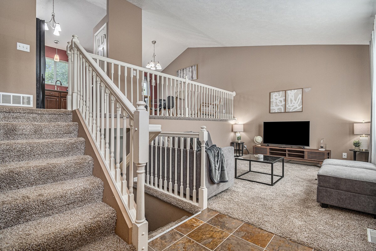 A spacious living area showcases a light-colored carpet and beige walls. A staircase leads to an upper level, with a railing visible. A television is mounted on the wall opposite a gray couch and a coffee table. Natural light filters in through large windows.