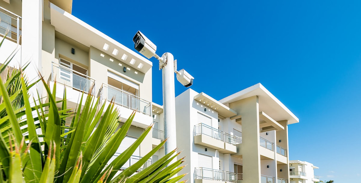 A modern residential complex is presented against a clear blue sky. Security cameras are mounted for safety, while palm leaves are visible in the foreground. The structure features balconies and large windows, contributing to a bright and inviting exterior.