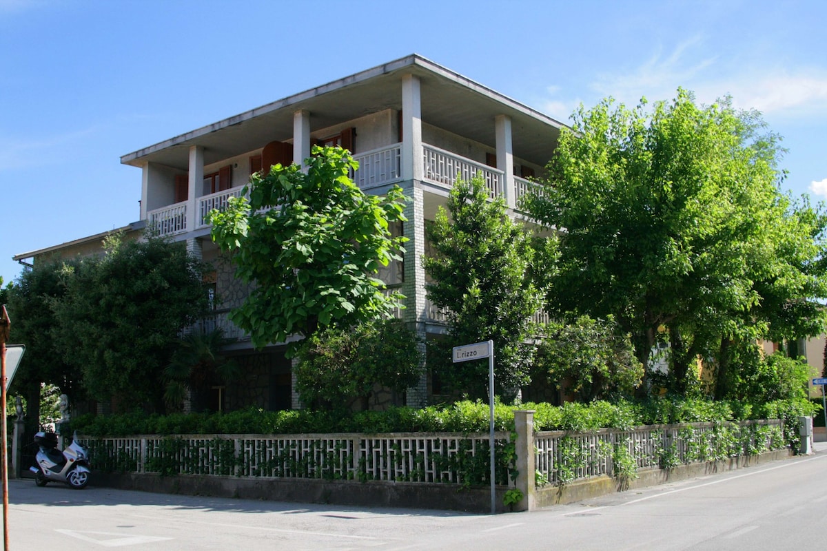 A three-story building is surrounded by lush greenery, featuring a balcony on the upper floor that overlooks the street. The facade is complemented by large windows, allowing for natural light. A low stone wall borders the property, providing a sense of privacy.