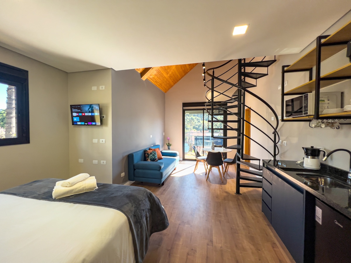 A modern studio apartment features a spacious bed and a cozy seating area with a blue sofa. A spiral staircase leads to an upper level. Natural light fills the room from large windows, and a kitchenette is equipped for preparing meals.