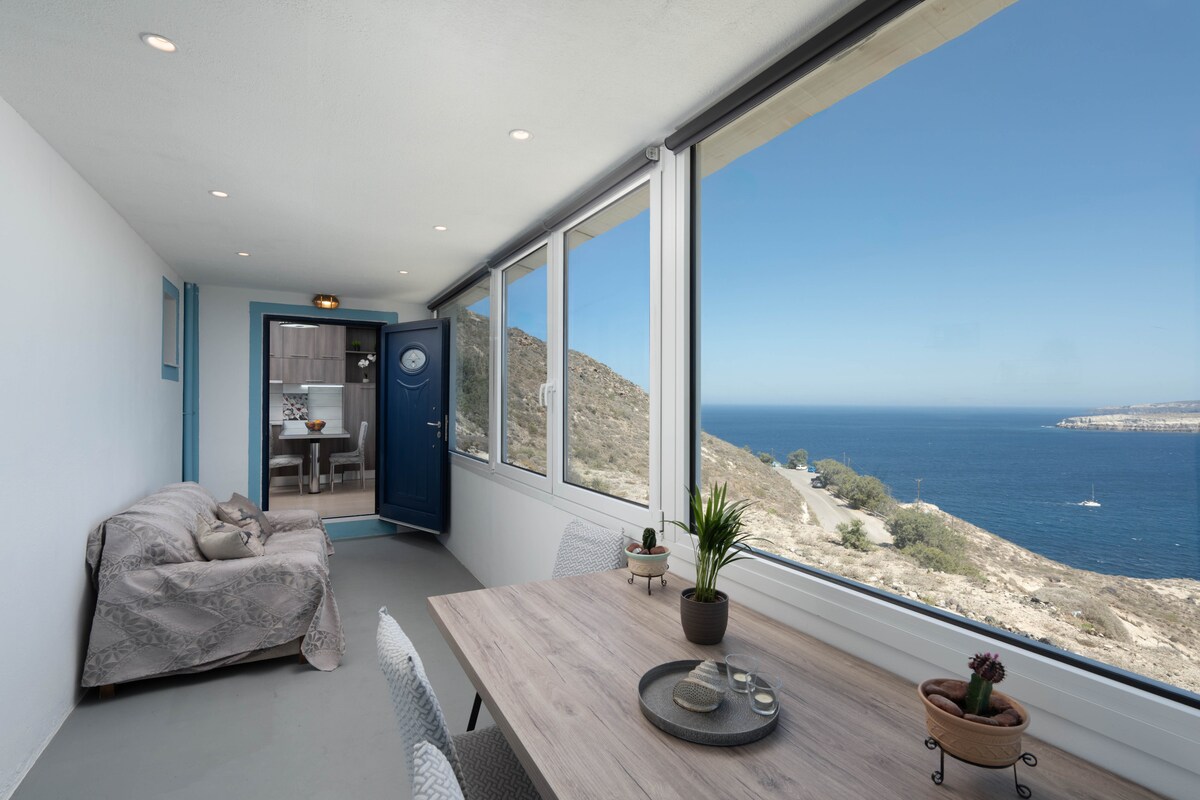 A bright enclosed veranda features a large table set for dining, complemented by a cozy sofa. Expansive windows provide unobstructed views of the sea and surrounding landscape, allowing natural light to fill the space.