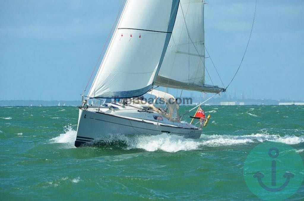 A sailboat is seen navigating through waves, with its white sails fully extended against a backdrop of blue sky. The vessel is depicted slicing through the water, illustrating its agility and speed in a lively maritime setting.