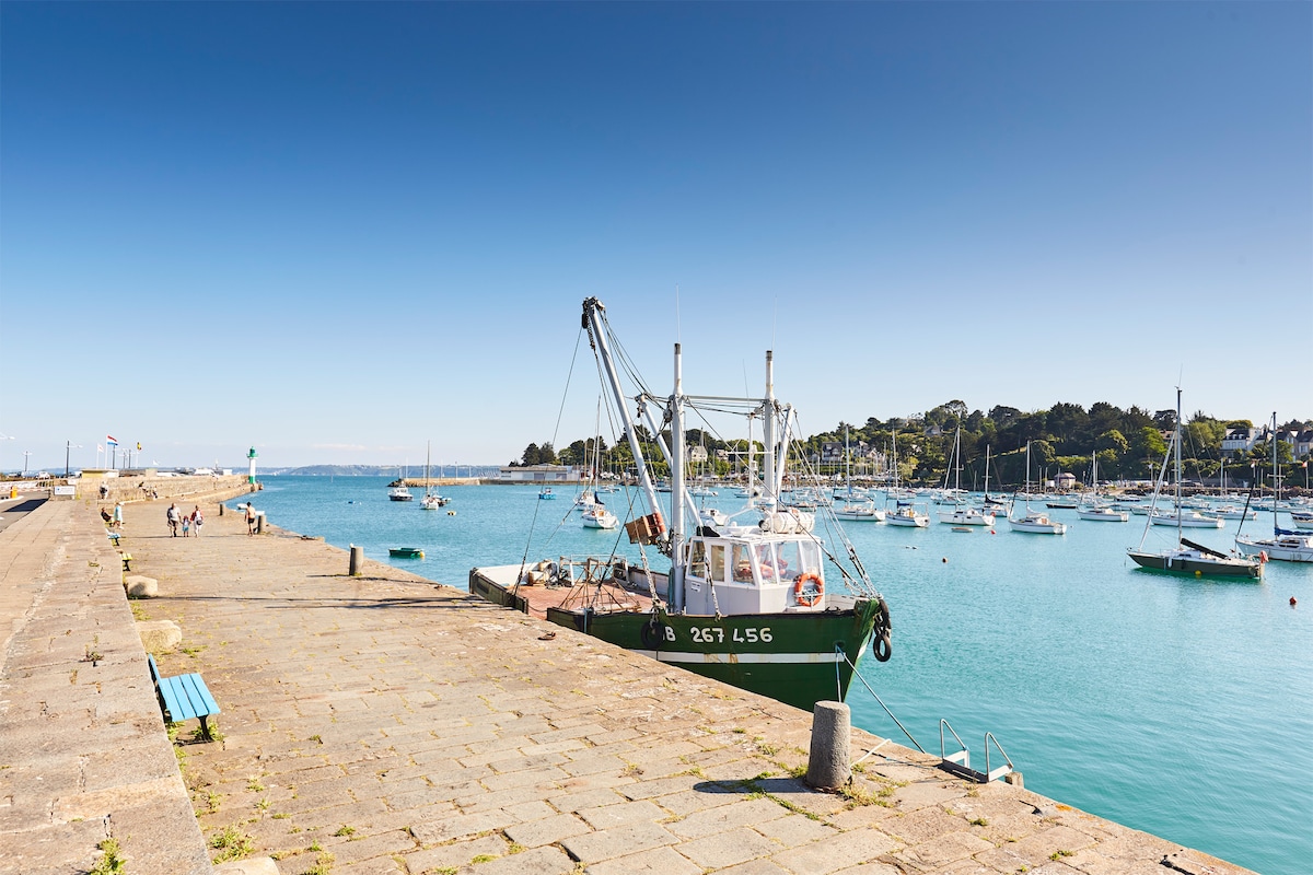 A fishing boat is docked at a stone pier, surrounded by a calm turquoise sea. Nearby, other boats are anchored, while the landscape is dotted with greenery and coastal buildings. Clear blue skies are seen above, providing a serene backdrop to the scene.