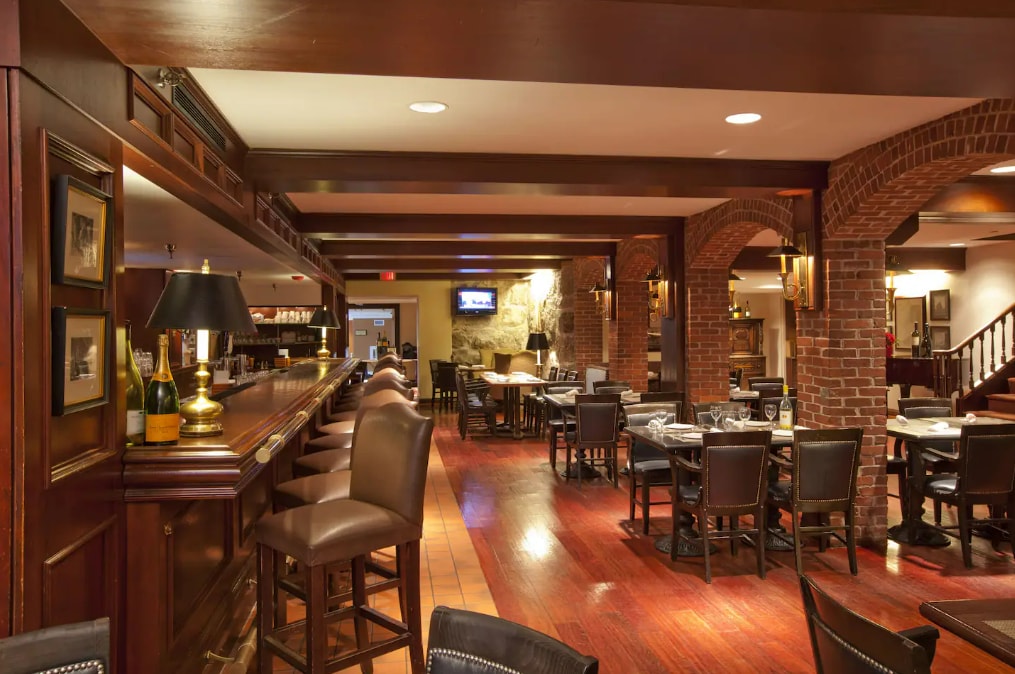 A spacious dining area features wooden beams and warm-toned brick accents. Dark wooden tables and chairs are arranged throughout. A well-lit bar with high stools is present, showcasing bottles and glassware. Soft lighting creates a relaxed atmosphere, and a staircase is visible in the background.