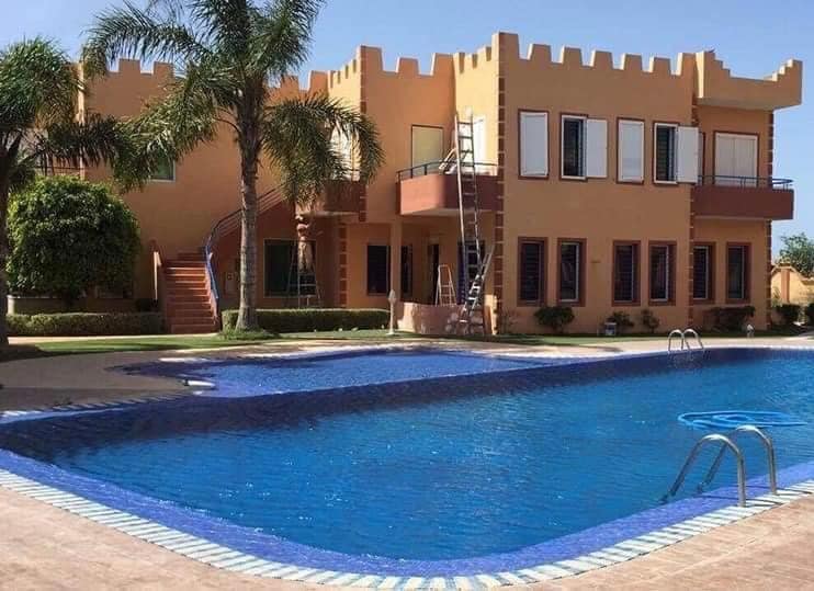 The exterior of the duplex features a castle-inspired design with a vibrant beige facade and multiple large windows. A clear blue pool is situated in the foreground, with surrounding palm trees and well-maintained landscaping, providing a serene outdoor environment.