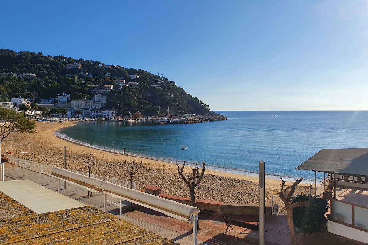 A serene beach scene captures a sandy shoreline curving around a tranquil bay. Gentle waves lap at the shore, with a view of greenery-covered hills in the background. A few boats can be seen in the water, adding to the peaceful ambiance of the location.