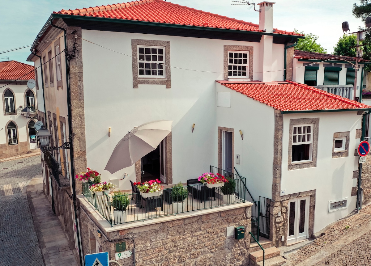 The exterior of the house features traditional stone walls and a warm terracotta roof. A spacious terrace is adorned with potted flowers and an umbrella, providing an inviting outdoor space. Nearby, cobblestone streets and neighboring buildings create a charming village atmosphere.