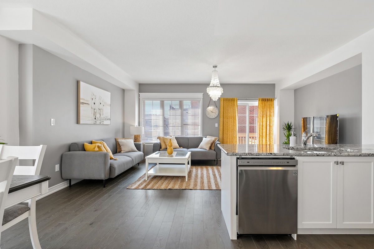 An open-concept living space is depicted, featuring sleek gray walls and a modern layout. A cozy sofa sits opposite a stylish coffee table, with large windows adorned with yellow curtains inviting natural light. The kitchen boasts contemporary appliances and elegant countertops, enhancing the inviting atmosphere.