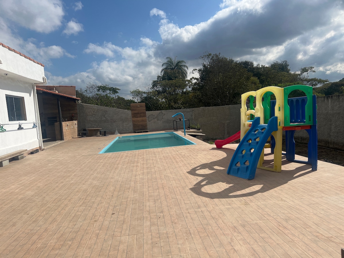A well-maintained outdoor space features a swimming pool surrounded by a tiled deck. Brightly colored play equipment, including a slide and climbing structure, is positioned nearby, offering a playful area for children. Lush greenery can be seen in the background, enhancing the setting.