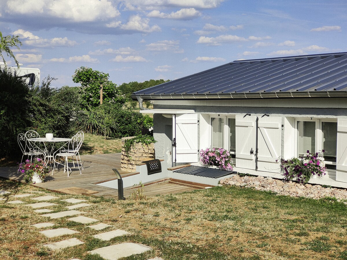 An outdoor seating area is visible, featuring a round table with two chairs surrounded by a grassy landscape. The exterior of the house displays decorative window shutters and potted flowers. A pathway of rectangular stones leads to the entrance, with trees and shrubs in the background.
