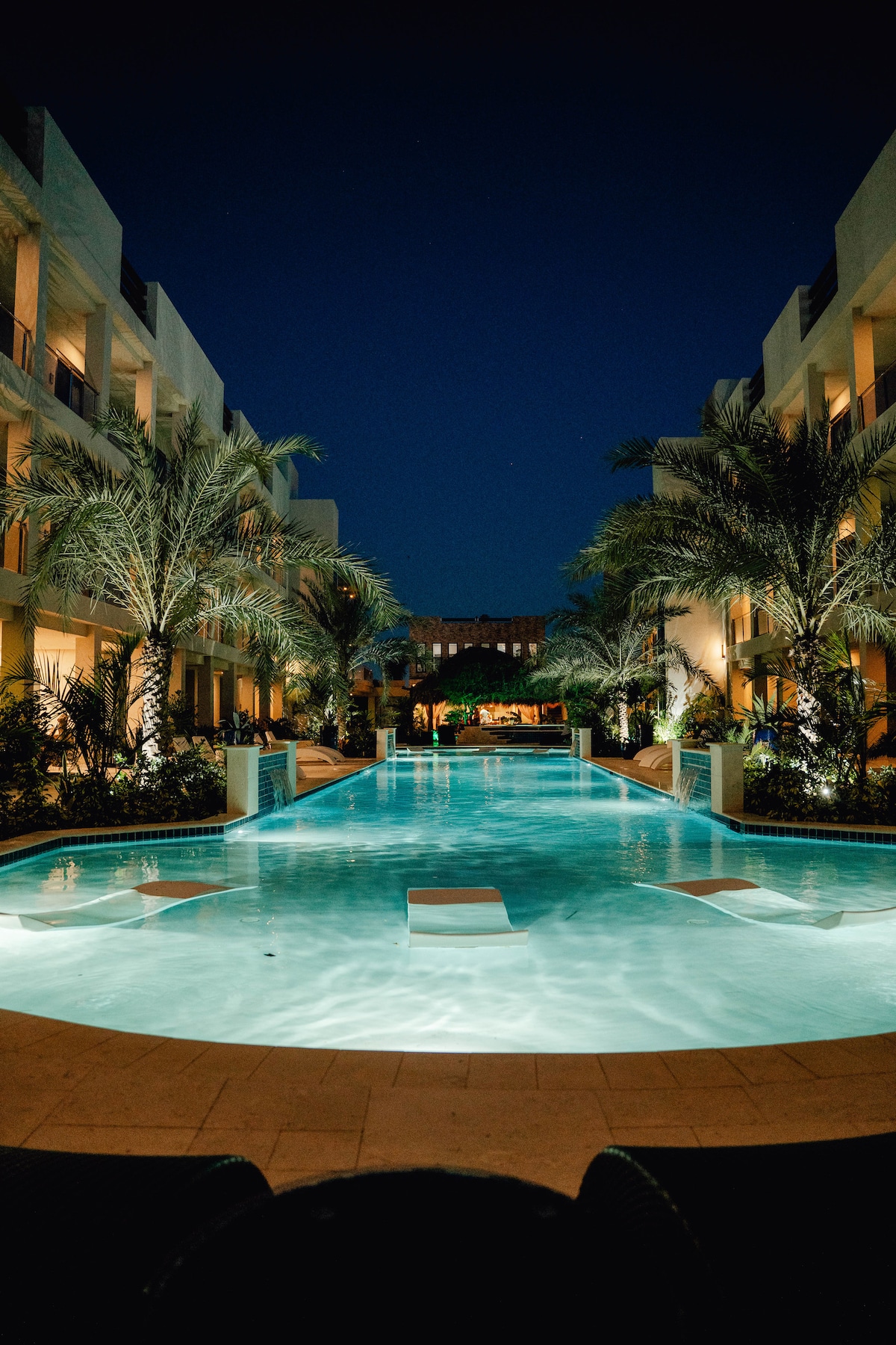A serene nighttime view of the pool area is captured, illuminated by soft lighting. Lush palm trees flank the edges, reflecting the tranquility of the space. The pool’s water glimmers under the night sky, creating a calm atmosphere amidst the surrounding modern buildings.