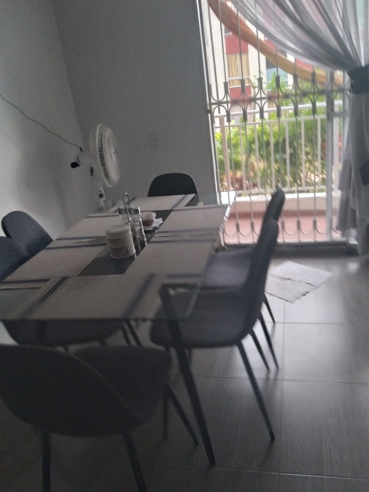 A dining area features a large glass table surrounded by several cushioned chairs. Natural light enters through a window adorned with light curtains, revealing a view of greenery outside. A small fan is positioned nearby, providing added comfort.