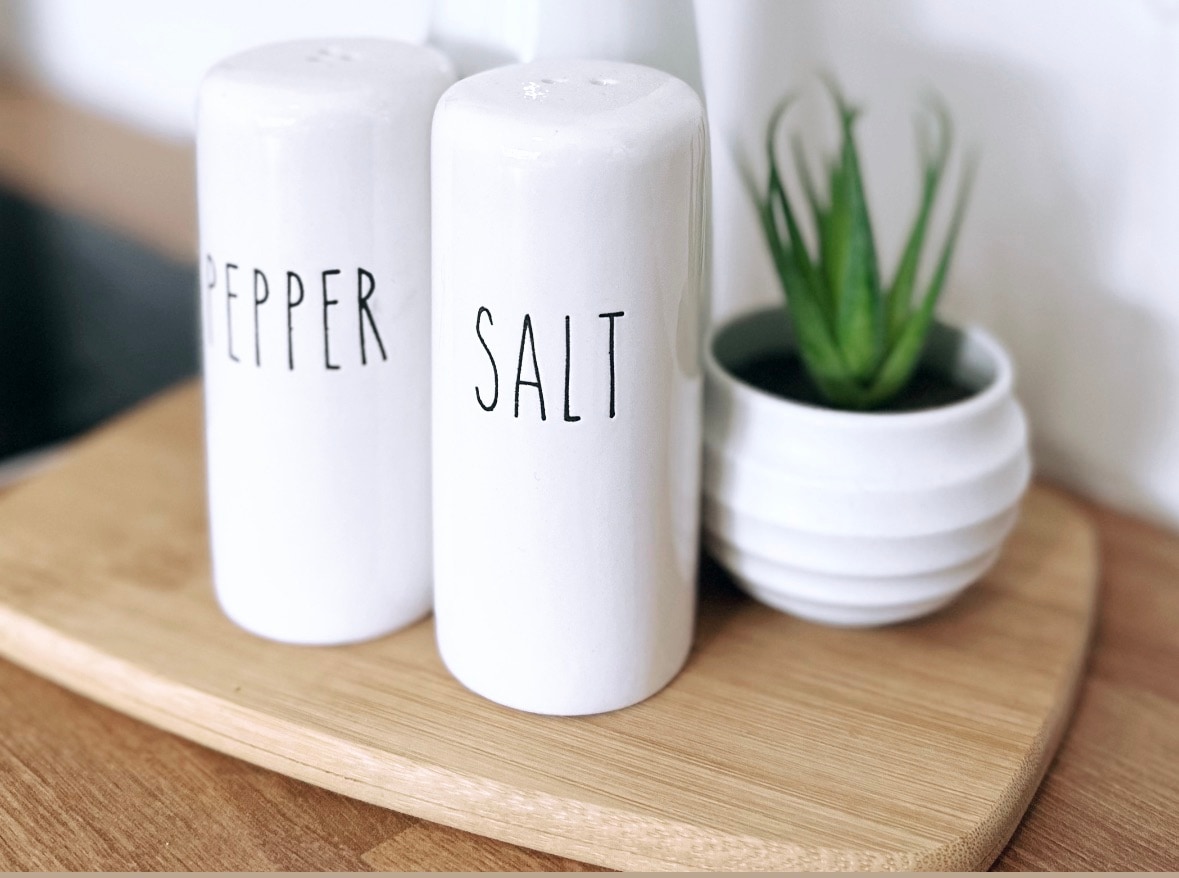 A wooden serving tray holds two white ceramic salt and pepper shakers, labeled clearly. A small potted plant with spiky leaves is positioned next to a white bowl, adding a subtle touch of greenery to the kitchen space.
