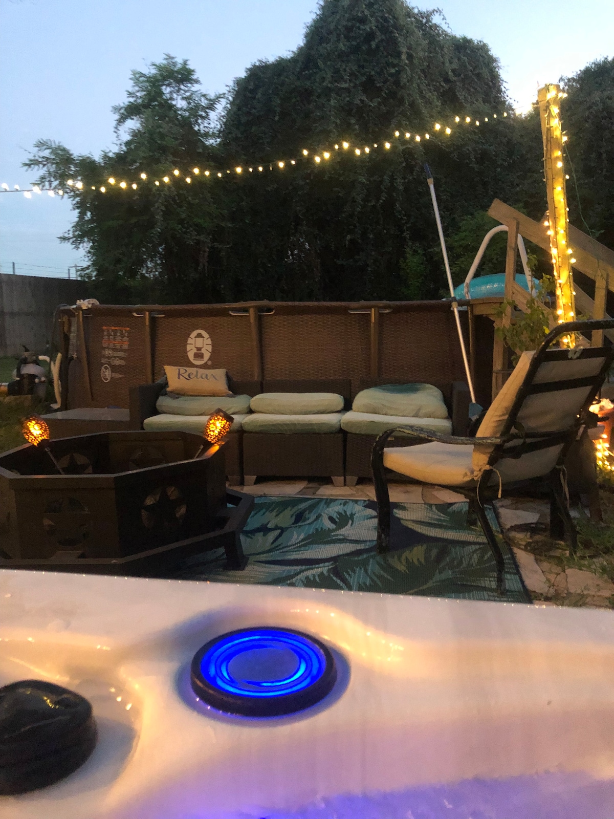 A cozy outdoor space is illuminated by soft string lights, featuring a comfortable seating area with plush cushions and a fire pit. A blue-lit hot tub is in the foreground, while a decorative rug adds a pop of color to the ground.