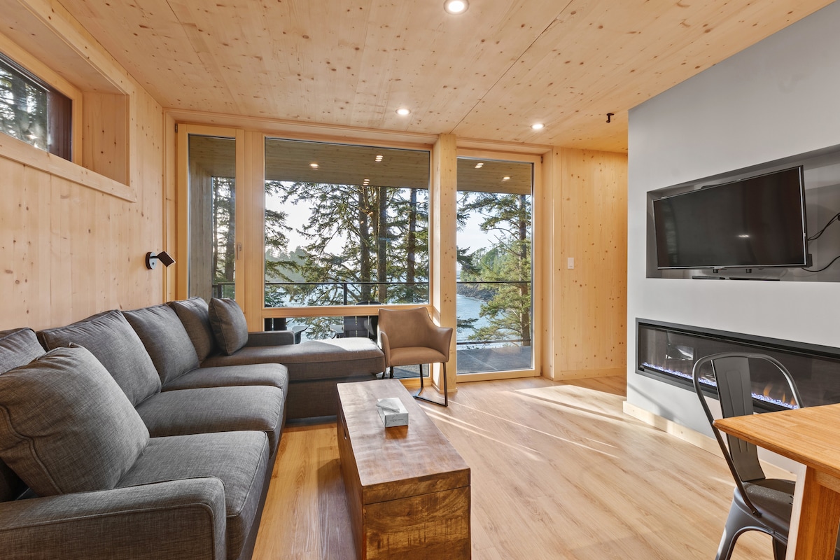 A comfortable living area features a large sectional sofa positioned near expansive windows, allowing views of the surrounding trees. A sleek wall-mounted TV is present, alongside an inviting fireplace. A wooden coffee table and a dining area with chairs complete the space, emphasizing a cozy atmosphere.