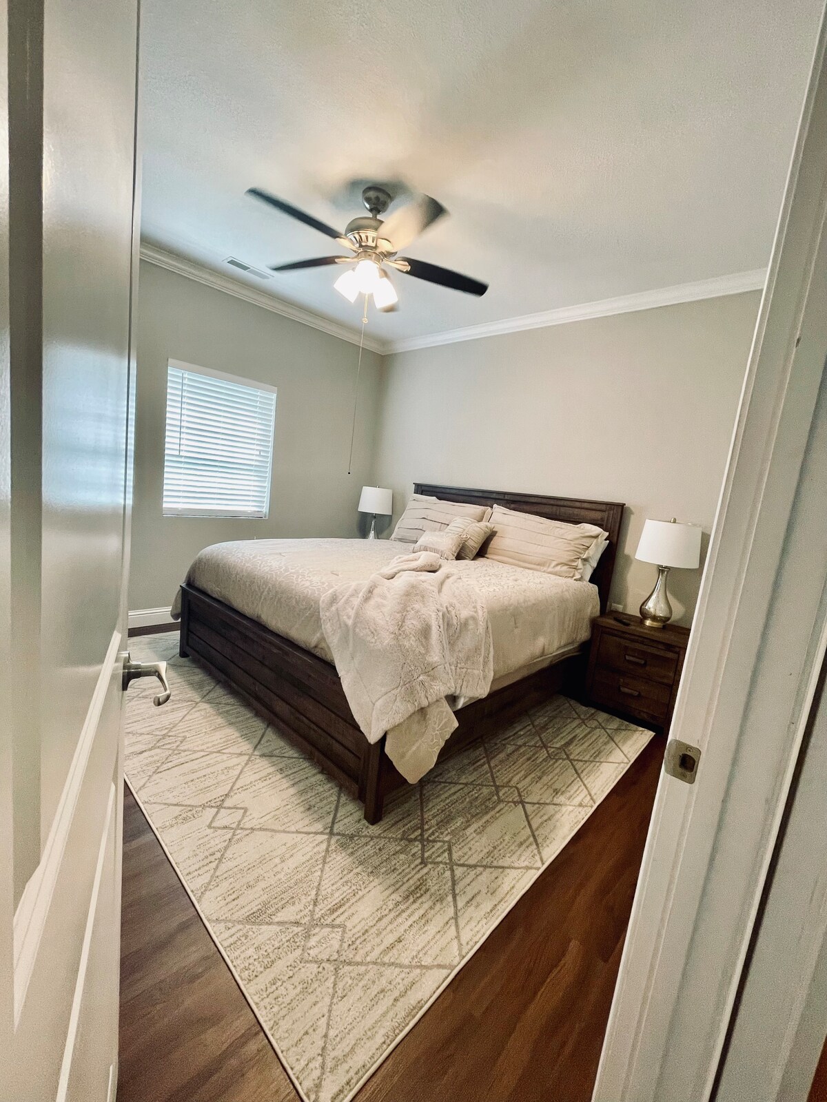 A serene bedroom is featured with a spacious bed, complemented by neutral bedding. Two bedside lamps are positioned on matching nightstands. A textured area rug lies beneath the bed, and a window with blinds allows natural light to gently illuminate the room.