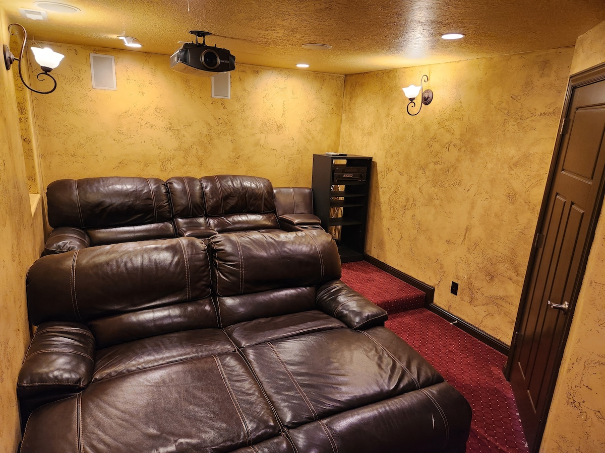 A dedicated theater room features luxury leather lounge couches arranged for comfort. Surrounding walls are painted in warm gold tones, creating a cozy atmosphere. A projector and audio speakers are installed for enhanced viewing experiences, while stairs lead to a slightly elevated area.