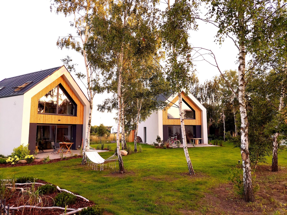 Two modern chalets are positioned side by side under the open sky, featuring large windows with wooden frames. Nearby, a hammock hangs between trees on a well-maintained lawn. Light reflects off the chalets' facades, showcasing their distinct architectural style against the natural backdrop.