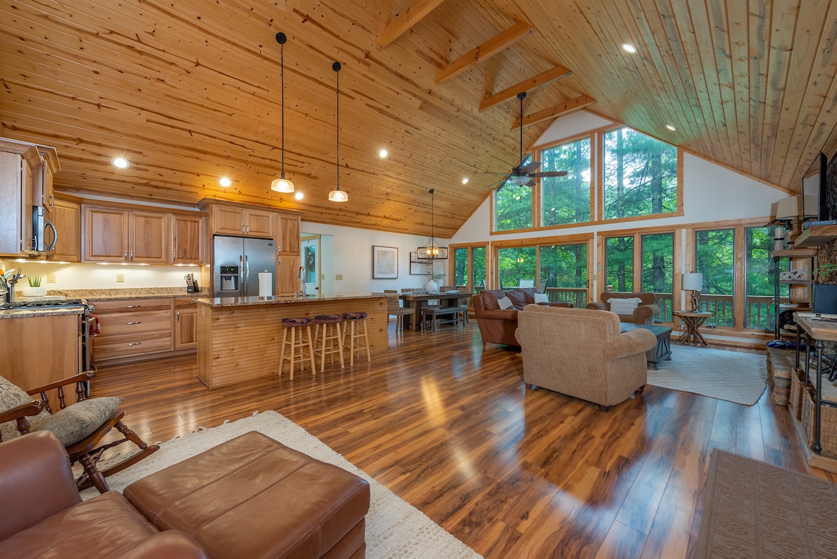 An open-concept living area showcases warm wood tones and high ceilings. A fully equipped kitchen features wooden cabinetry and a granite countertop. Comfortable seating is arranged near large windows, providing ample natural light and a view of the surrounding greenery.