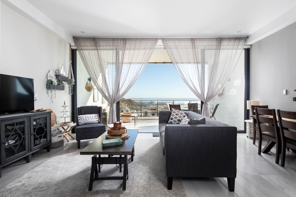 A spacious living area is revealed, featuring large glass doors that open to stunning views. Sheer drapery softly filters light, while a comfortable sofa and dark wood furniture create a welcoming ambiance. A flat-screen television is positioned beside a decorative cabinet.