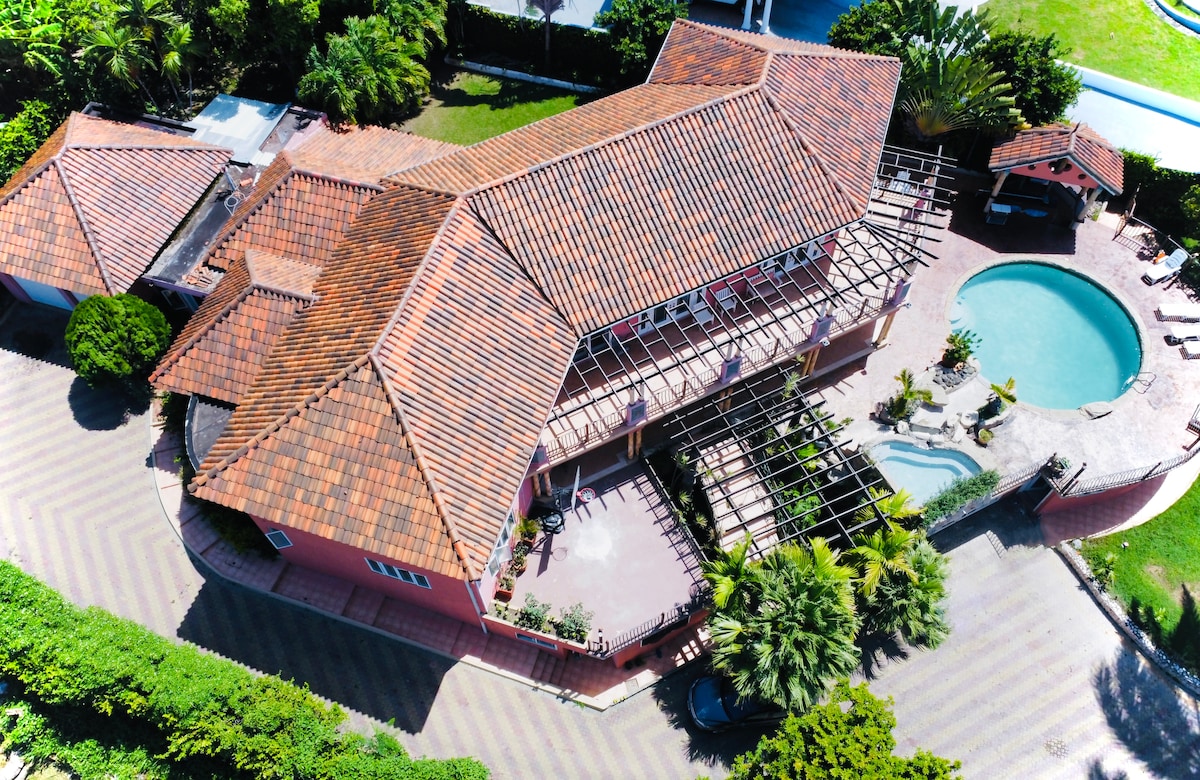 Aerial view of a villa with a distinctive terracotta roof and surrounding lush greenery. The layout includes a private pool and landscaped areas. A pergola structure extends from the villa, enhancing outdoor space. The overall setting reflects a serene and spacious environment.